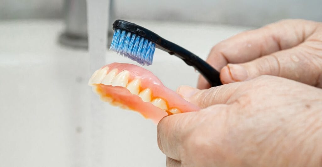Jak dbać o protezę zębową - starsza osoba myjąca protezę zębową szczoteczką pod kranem